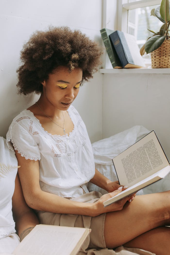 A young woman with natural hair enjoys reading a book by the window with warm sunlight.